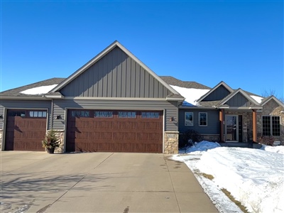 A modern single-story home with a gray exterior, multiple garages, and snow patches on the driveway under a clear blue sky.