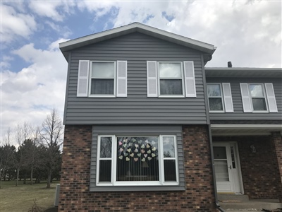 A two-story home with gray siding and brick accents, featuring decorative window shutters and a cloudy sky backdrop.