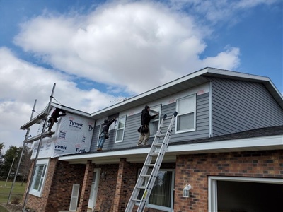 Two workers on ladders installing siding on a two-story house with gray and brick exterior under a cloudy sky.