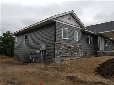 A house under construction with gray siding and stone accents, featuring a dirt yard and overcast sky.