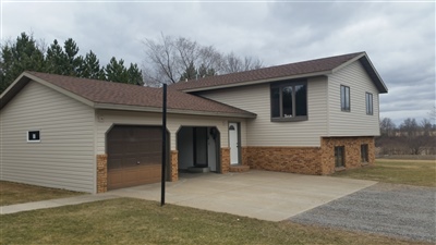A split-level home with beige siding and brick accents, featuring a single garage and a gravel driveway, set against a cloudy sky.