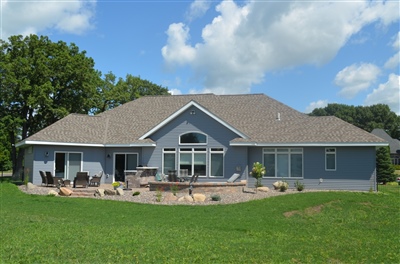 A spacious single-story home with a blue facade, featuring large windows and a patio area, set in a lush green yard under a clear sky.