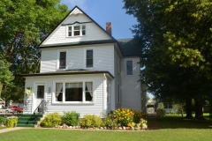 A charming white two-story house with green trim, surrounded by lush greenery and a well-manicured garden under a sunny sky.