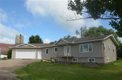 A beige single-story house with a double garage and a patio, set on a landscaped lawn with a few trees under a cloudy sky.