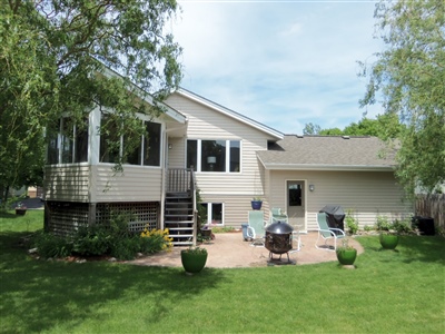  A split-level home with beige siding, featuring a deck and landscaped backyard with green lawn and trees under a sunny sky.