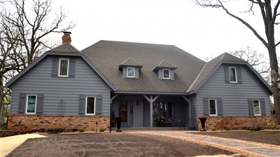  A large gray house with symmetrical gabled roofs, featuring a central entryway and circular driveway, set against a backdrop of bare trees.