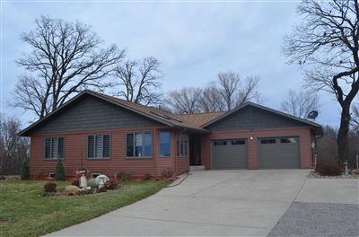 A red brick single-story house with a double garage and concrete driveway, surrounded by leafless trees under an overcast sky.