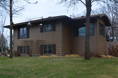 A dark brown two-story house with large windows, nestled among bare trees and a leaf-covered lawn under an overcast sky.