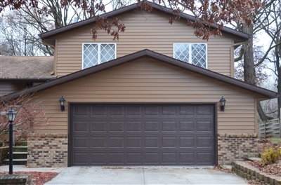 A brown two-story home with an oversized garage door, large upper windows, and surrounding leafless trees against an overcast sky.