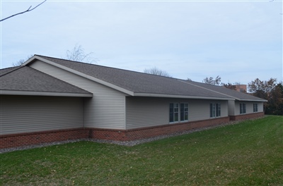 A long, beige single-story building with a sloped roof and multiple windows, set against a grassy backdrop under a cloudy sky.