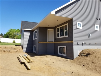 A house under construction with gray siding and brown trim, featuring an unfinished yard and exposed dirt, under a clear blue sky.