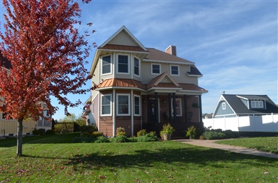 A traditional two-story house with a brick and beige facade, featuring a vibrant red tree in the front yard under a clear blue sky.