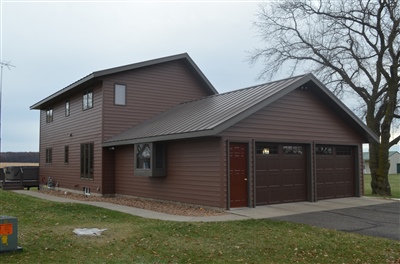 A dark brown two-story home with a large attached garage, set against a bare tree line under an overcast sky.