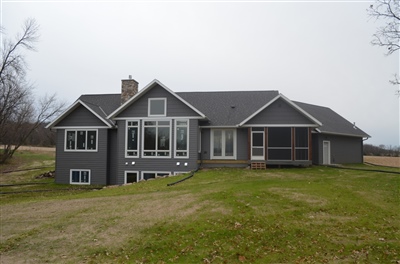 A modern gray single-story house with large windows and a sprawling layout, set on a grassy field under a cloudy sky.