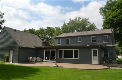 A modern gray two-story home with a large patio area, set against a backdrop of lush green trees and a well-manicured lawn.