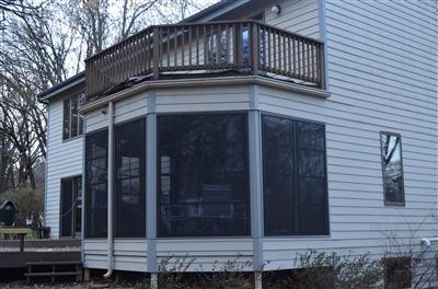 A white house with a screened-in porch on the lower level and a wooden deck above, surrounded by trees.