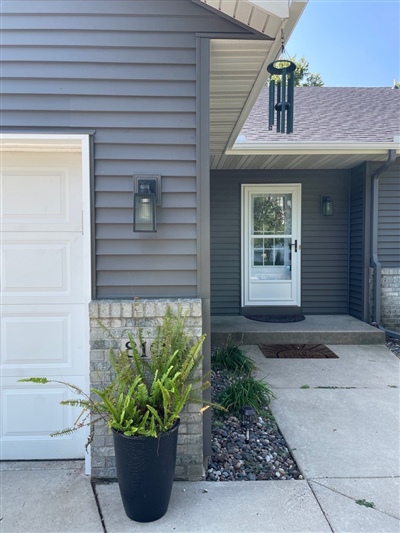 A close-up of a house entrance featuring gray siding, a modern glass door, and a decorative plant, under a clear blue sky.
