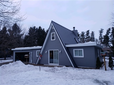 A unique A-frame house with gray siding and an attached garage, surrounded by snow-covered ground and bare trees.