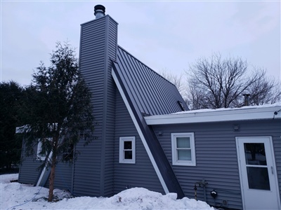 A gray A-frame house with a tall chimney, featuring a snow-covered roof and surrounding landscape under a cloudy winter sky.