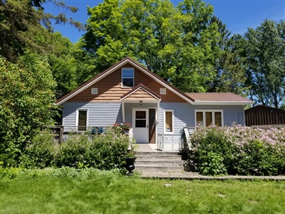 A quaint house with beige and brown siding, featuring a central entrance and surrounded by lush greenery and mature trees under a clear blue sky.