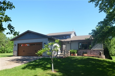  A modern split-level house with beige siding and a red garage door, surrounded by lush greenery and a young tree under a clear blue sky.