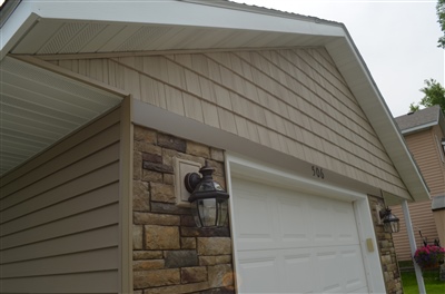 A close view of a beige home with stone accents, showing a worker on a ladder next to the roof and a garage door.