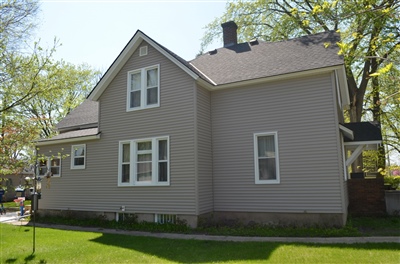  Side view of a two-story taupe house with white trim, surrounded by a well-maintained lawn and trees on a sunny day.