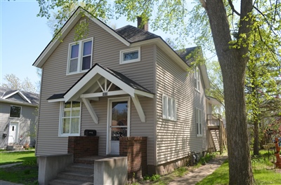 Front view of a two-story taupe house with a small porch, white trim, and gable roof, surrounded by trees on a sunny day.