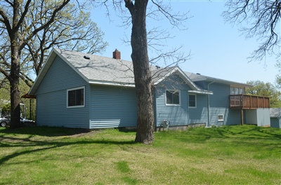 Side view of a single-story blue house with a spacious backyard, deck, and mature trees on a bright, sunny day.