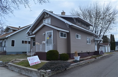 Small taupe house with white trim and front porch, located on a corner lot with a yard sign, on an overcast day.