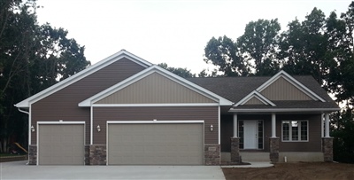 Newly constructed single-story home with a three-car garage, brown and tan exterior, stone accents, and surrounded by trees.