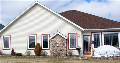  A modern single-story house with beige siding and red window trim, featuring a stone accent section near the entrance and a snowy front yard.