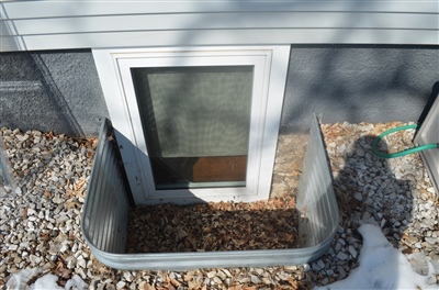 A close-up of a house foundation vent, surrounded by a frame filled with small stones and placed against a gray siding background.