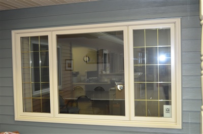 A large three-pane window with white trim, providing a clear view into a room illuminated by interior lighting, set against gray siding.