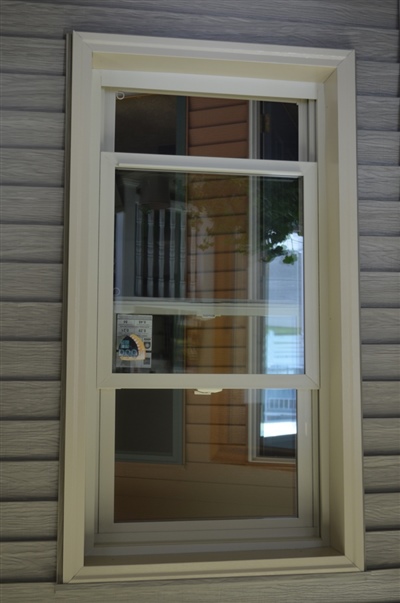 A tall vertical window with white trim, providing a multi-layered view of books on a shelf inside, set against a background of gray siding.