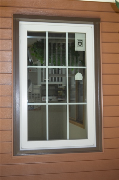  A tall vertical window with white trim and grid design, offering a glimpse into an indoor space against a backdrop of reddish-brown siding.