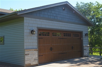 Close-up of a garage with light blue siding, stone accents, and a large wooden garage door, surrounded by greenery on a sunny day.