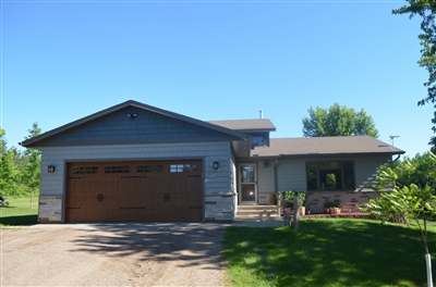 A contemporary single-story home with gray siding and a brown garage door, featuring lush green landscaping under a clear blue sky.