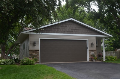 A garage facade of a house with beige siding and a large brown door, surrounded by lush greenery and trees.