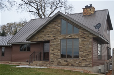 A distinctive house with stone walls and large windows, featuring a metal roof and a chimney, set against a backdrop of bare trees.