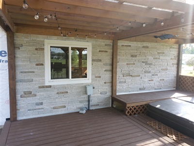 A covered deck area with light-colored stone walls, wooden ceiling, and a large window, connected to a dark brown wooden floor.