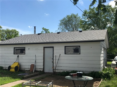 A small white house with a gray shingled roof, a central door, and two small windows, with garden tools and equipment outside.