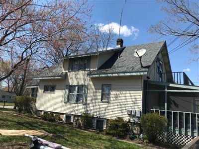 A beige house with a gray shingled roof, an attic window, a satellite dish, and a side porch, surrounded by a yard with trees and bushes.