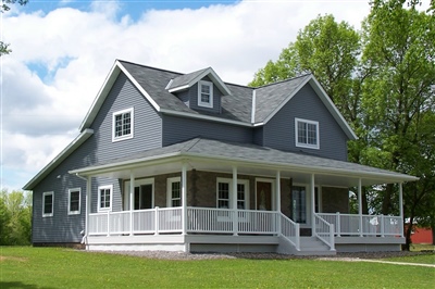  A gray house with a white wraparound porch, gable roof, dormer windows, and surrounded by a green yard with trees in the background.