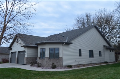 A beige house with a gray roof, featuring a three-car garage, a bay window, and a paved driveway, surrounded by a yard with bare trees.