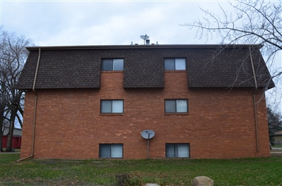 A three-story red brick apartment building with a dark brown mansard roof, multiple windows, and a satellite dish on the front facade.