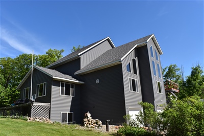 A modern dark gray house with a steep gabled roof, multiple windows, and surrounded by a green yard and trees.